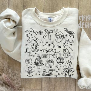 May include: Cream-colored sweatshirt with a black line drawing of Christmas-themed icons, including a gingerbread man, Santa, and a Christmas tree. The words "merry christmas" are written in the center of the design. The sleeves are rolled up.