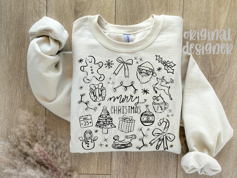 May include: Cream-colored sweatshirt with a black line drawing of Christmas-themed icons, including a gingerbread man, Santa, and a Christmas tree. The words "merry christmas" are written in the center of the design. The sleeves are rolled up.