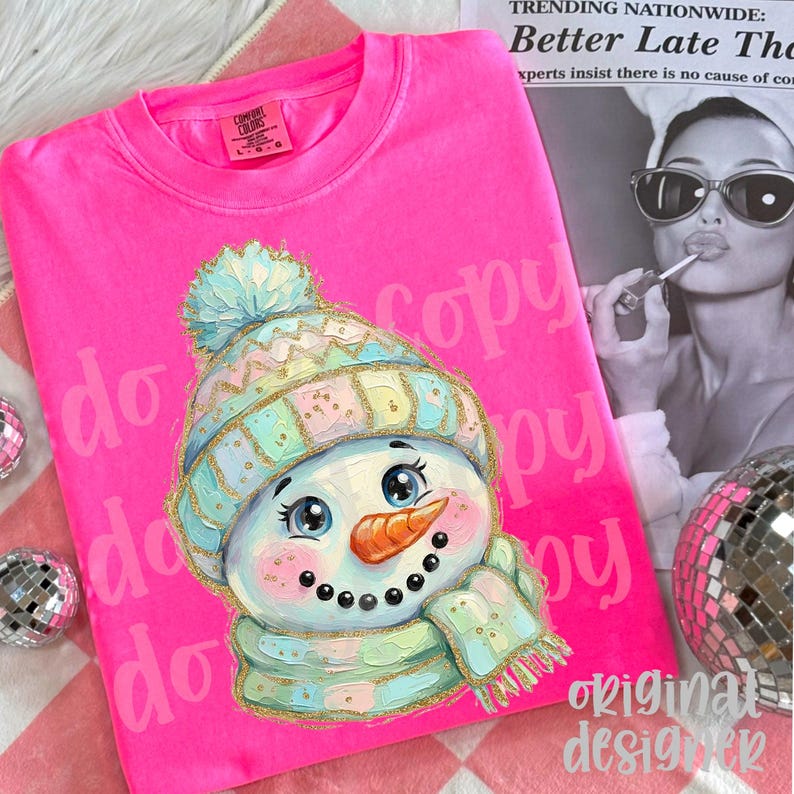 May include: A bright neon pink t-shirt featuring a graphic illustration of a snowman wearing a pastel-colored knit hat and scarf with gold glitter accents. The snowman has a carrot nose, black button eyes, and a smiling mouth.