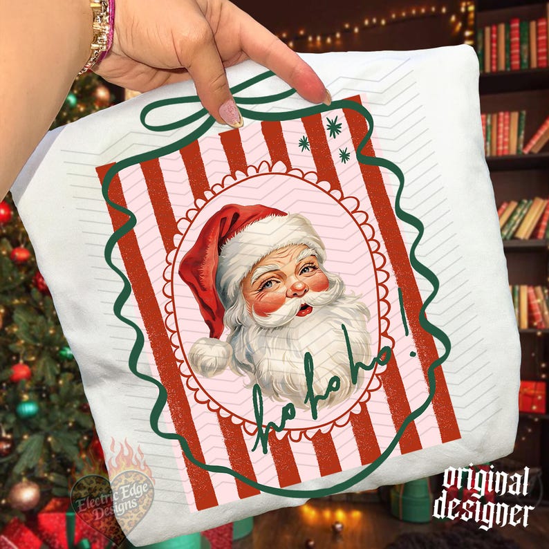 May include: A white sweatshirt featuring a graphic of Santa Claus wearing a red hat inside a pink scalloped oval frame. The background has red and white vertical stripes with a dark green ribbon border and the text ho ho ho in green script.