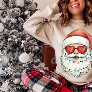 May include: A beige sweatshirt featuring a Santa Claus graphic with heart-shaped sunglasses and a string of colorful lights around his beard. The person is wearing red and black plaid pajama pants. A decorated Christmas tree is in the background.
