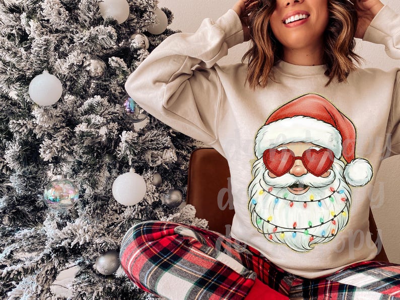 May include: A beige sweatshirt featuring a Santa Claus graphic with heart-shaped sunglasses and a string of colorful lights around his beard. The person is wearing red and black plaid pajama pants. A decorated Christmas tree is in the background.