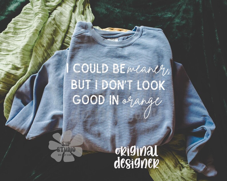 May include: A blue sweatshirt with the text "I COULD BE meaner, BUT I DON'T LOOK GOOD IN orange" in white. The sweatshirt is folded on a green and dark green background. The words "SIP STUDIO" and "original designer" are also visible.