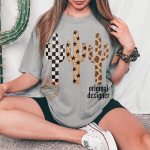 Może przedstawiać: Szara koszulka z trzema wzorami kaktus&oacute;w w r&oacute;żnych wzorach: w szachownicę, w gwiazdy i w krowią sk&oacute;rę. Tekst "original designer" jest wydrukowany pod kaktusami. Koszulka ma luźny kr&oacute;j.
