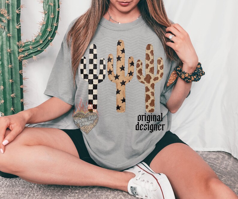 Może przedstawiać: Szara koszulka z trzema wzorami kaktus&oacute;w w r&oacute;żnych wzorach: w szachownicę, w gwiazdy i w krowią sk&oacute;rę. Tekst "original designer" jest wydrukowany pod kaktusami. Koszulka ma luźny kr&oacute;j.
