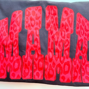 May include: A black sweatshirt featuring the word MAMA in large, bold, block letters. The letters are filled with a vibrant red and pink leopard print pattern that stands out against the dark fabric of the garment, creating a high-contrast design.
