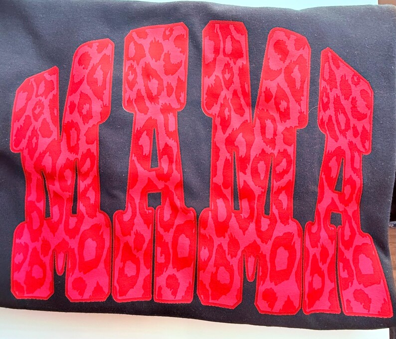 May include: A black sweatshirt featuring the word MAMA in large, bold, block letters. The letters are filled with a vibrant red and pink leopard print pattern that stands out against the dark fabric of the garment, creating a high-contrast design.