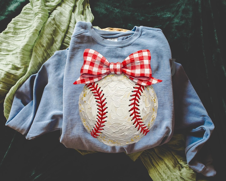 Peut inclure: Un sweat-shirt bleu clair avec un motif de baseball. La balle est blanche avec des coutures rouges et un n&oelig;ud vichy rouge et blanc. Le baseball a des d&eacute;tails dor&eacute;s.