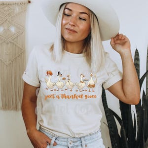 May include: Cream-colored t-shirt featuring a whimsical illustration of five geese, each adorned with festive hats and holding plates of food. The text "just a thankful goose" is printed below the geese in a warm, orange hue. The shirt is worn by a person.