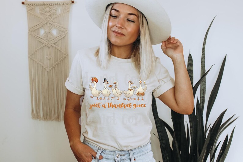 May include: Cream-colored t-shirt featuring a whimsical illustration of five geese, each adorned with festive hats and holding plates of food. The text "just a thankful goose" is printed below the geese in a warm, orange hue. The shirt is worn by a person.