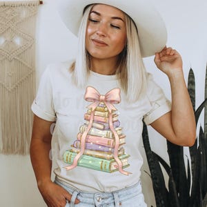 May include: Cream-colored t-shirt featuring a stack of books with a pink bow and ribbon design. The books are in pastel colors, and the text "Do More of What Makes You Happy" is visible on the shirt.