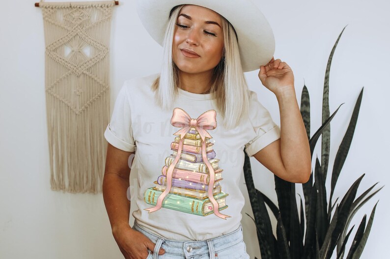 May include: Cream-colored t-shirt featuring a stack of books with a pink bow and ribbon design. The books are in pastel colors, and the text "Do More of What Makes You Happy" is visible on the shirt.