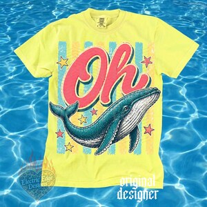May include: Yellow t-shirt with a graphic of a blue whale and the word "Oh" in red. The design includes stars and vertical stripes in blue, yellow, and pink. The shirt has a crew neck and short sleeves.