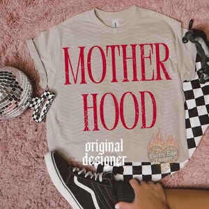 May include: A beige short-sleeved t-shirt featuring the red distressed text MOTHER HOOD printed on the front. The shirt has a subtle chevron pattern and includes a small heart-shaped logo with flames and the text Electric Edge Designs.