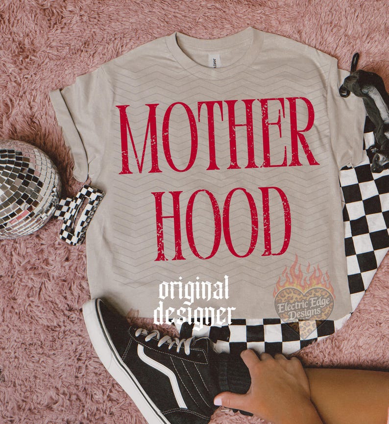 May include: A beige short-sleeved t-shirt featuring the red distressed text MOTHER HOOD printed on the front. The shirt has a subtle chevron pattern and includes a small heart-shaped logo with flames and the text Electric Edge Designs.