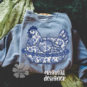 May include: A light blue sweatshirt featuring a detailed blue and white floral hen design. The hen is sitting on a decorative plate. The words "original designer" are printed in white script on the bottom right.
