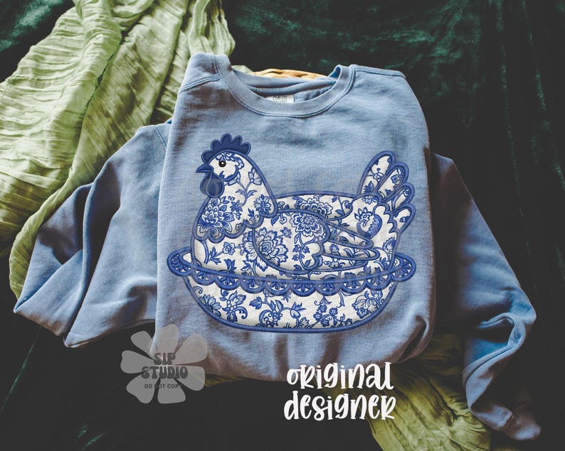May include: A light blue sweatshirt featuring a detailed blue and white floral hen design. The hen is sitting on a decorative plate. The words "original designer" are printed in white script on the bottom right.