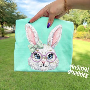 May include: A light turquoise t-shirt featuring a detailed illustration of a white bunny with pink-lined ears, wearing purple glasses and a green bow. The text "original designer" is printed on the lower right corner.