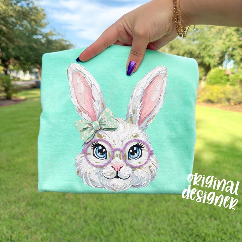 May include: A light turquoise t-shirt featuring a detailed illustration of a white bunny with pink-lined ears, wearing purple glasses and a green bow. The text "original designer" is printed on the lower right corner.
