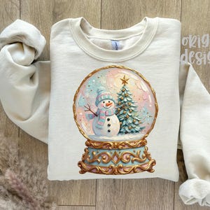 May include: A cream-coloured crewneck sweatshirt featuring a graphic of a snow globe containing a snowman wearing a blue and pink striped scarf and hat, next to a Christmas tree. The base of the globe is ornate gold and light blue.