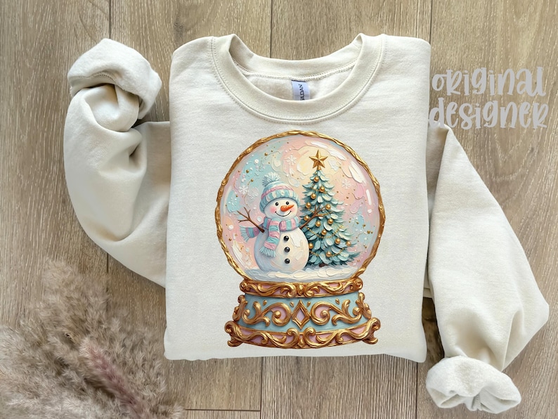 May include: A cream-coloured crewneck sweatshirt featuring a graphic of a snow globe containing a snowman wearing a blue and pink striped scarf and hat, next to a Christmas tree. The base of the globe is ornate gold and light blue.