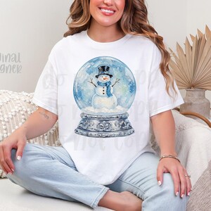 May include: White t-shirt featuring a watercolor illustration of a snowman inside a snow globe. The snowman wears a black top hat and blue scarf. The snow globe sits on a decorative base. The words "original designer" are printed on the shirt.