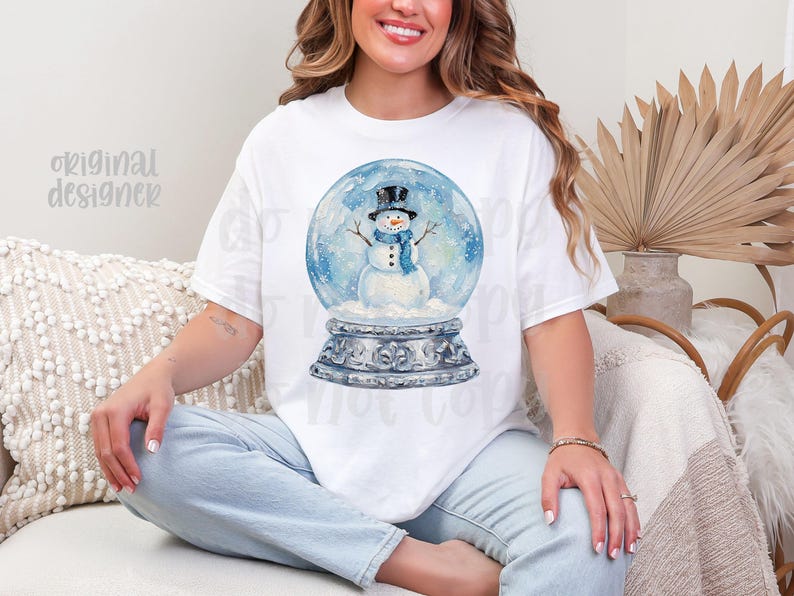May include: White t-shirt featuring a watercolor illustration of a snowman inside a snow globe. The snowman wears a black top hat and blue scarf. The snow globe sits on a decorative base. The words "original designer" are printed on the shirt.