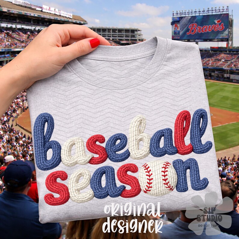 May include: A light gray t-shirt with the words "Baseball Season" in blue, red, and white, with a baseball graphic. The shirt is held up in front of a baseball stadium.
