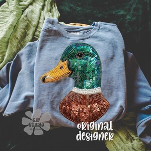 Pu&ograve; includere: Una felpa azzurra con un design dettagliato di un'anatra. La testa dell'anatra &egrave; impreziosita da paillettes verdi, un becco di perline giallo e un collo di perline marroni. Le parole "original designer" sono stampate in basso a destra.