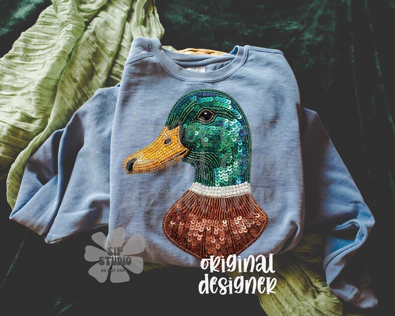 Pu&ograve; includere: Una felpa azzurra con un design dettagliato di un'anatra. La testa dell'anatra &egrave; impreziosita da paillettes verdi, un becco di perline giallo e un collo di perline marroni. Le parole "original designer" sono stampate in basso a destra.
