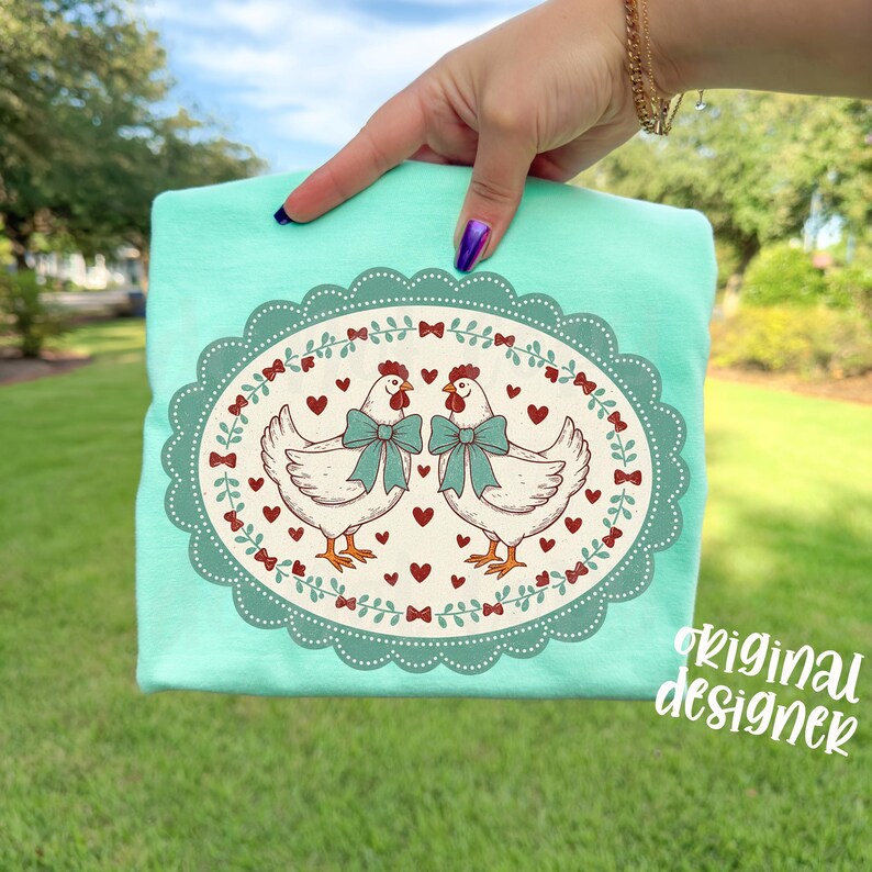 May include: A light teal t-shirt featuring an oval design with two white chickens wearing teal bows. The design includes red hearts and bows, with the text "original designer" in a script font.