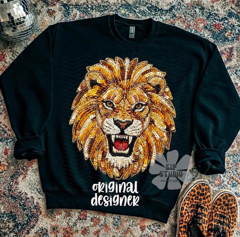 May include: Black sweatshirt featuring a detailed, sequined lion's head design in gold, orange, and white. The text "original designer" is printed in white script. Leopard print sneakers are visible in the bottom right corner.
