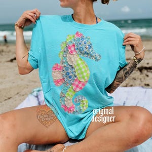 May include: A turquoise t-shirt featuring a colorful seahorse applique. The seahorse design incorporates floral, gingham, and lace fabrics, accented with pearls. The words "original designer" are printed on the shirt.