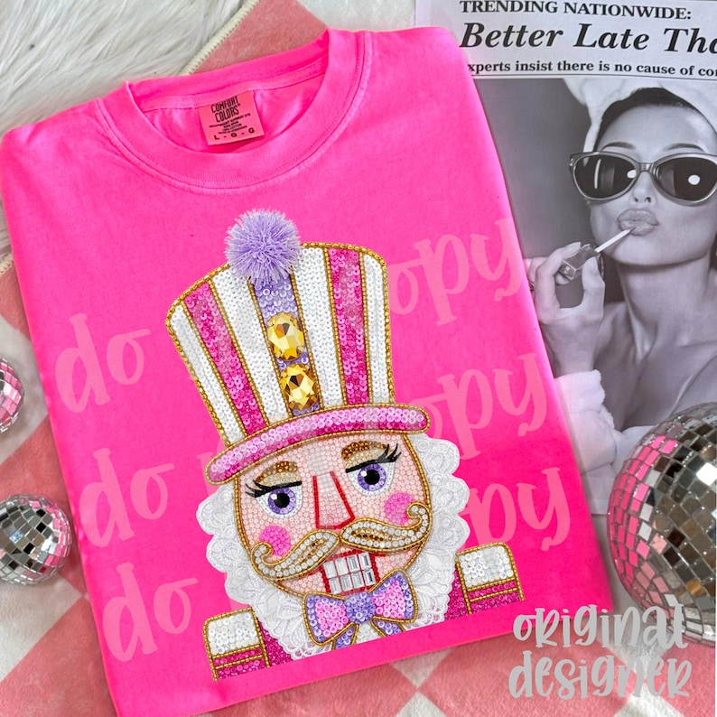 May include: Bright pink Comfort Colors sweatshirt featuring a detailed nutcracker design. The nutcracker has a sequined hat, a purple pom-pom, and a bow tie. The words "original designer" are at the bottom right.