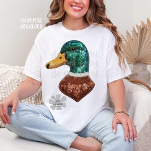 Pu&ograve; includere: T-shirt bianca con un disegno di anatra con paillettes. L'anatra ha la testa verde, il becco giallo e il corpo con paillettes marroni. Le parole "original designer" sono stampate in alto a sinistra sulla maglietta.