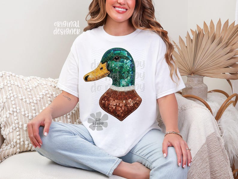 Pu&ograve; includere: T-shirt bianca con un disegno di anatra con paillettes. L'anatra ha la testa verde, il becco giallo e il corpo con paillettes marroni. Le parole "original designer" sono stampate in alto a sinistra sulla maglietta.