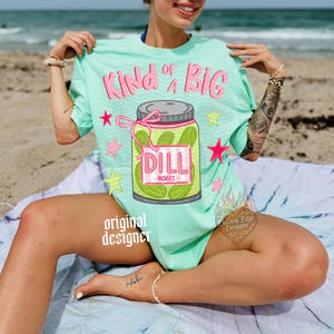 May include: A light green t-shirt with the text "Kind of a Big" in pink, and a jar of pickles graphic. The jar has a pink label that says "Dill Pickles". Pink stars and green stars are scattered around the graphic.