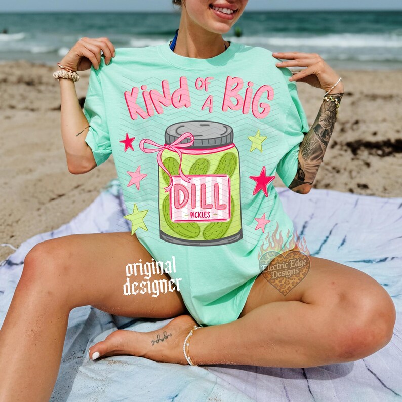 May include: A light green t-shirt with the text "Kind of a Big" in pink, and a jar of pickles graphic. The jar has a pink label that says "Dill Pickles". Pink stars and green stars are scattered around the graphic.