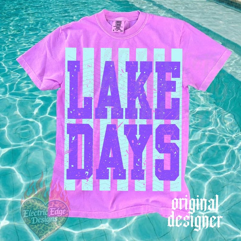 May include: A lilac-colored t-shirt with the words "LAKE DAYS" in purple block letters. The text is set against a backdrop of light blue vertical stripes. The shirt has a crew neck and short sleeves. The words "original designer" are printed on the bottom right.