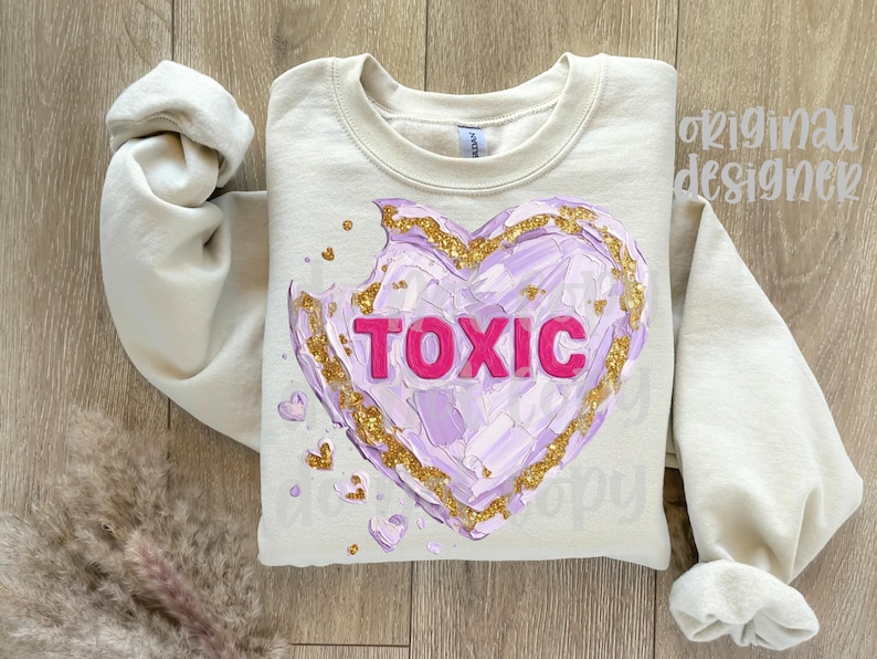 May include: A cream-colored sweatshirt featuring a large, textured heart design. The heart is primarily lavender with gold accents and the word "TOXIC" in bright pink. Small heart details are scattered around the main design.