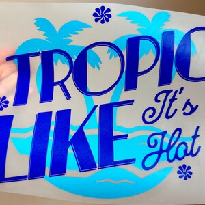 May include: A heat transfer decal on a clear backing sheet featuring the dark blue text TROPIC LIKE It's Hot. The design includes a light blue silhouette of palm trees and ocean waves, accented by three small dark blue flower icons.