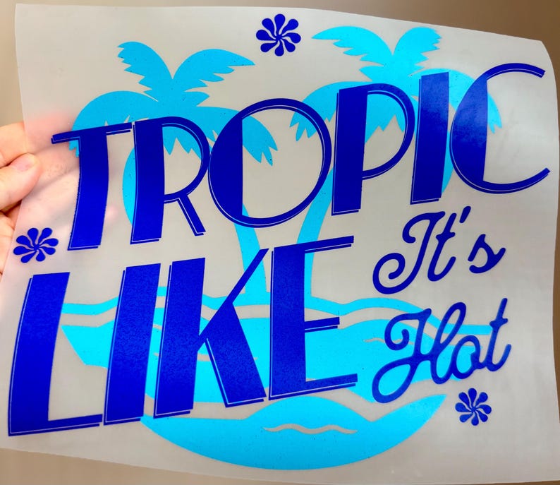May include: A heat transfer decal on a clear backing sheet featuring the dark blue text TROPIC LIKE It's Hot. The design includes a light blue silhouette of palm trees and ocean waves, accented by three small dark blue flower icons.