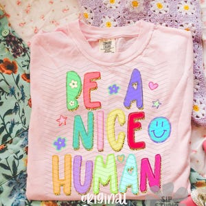 May include: A light pink Comfort Colors t-shirt with the phrase "BE A NICE HUMAN" in colorful, glitter-outlined letters. The design includes stars, flowers, and a smiley face. The words "original designer" and "SIP STUDIO DO NOT COPY" are also printed on the shirt.