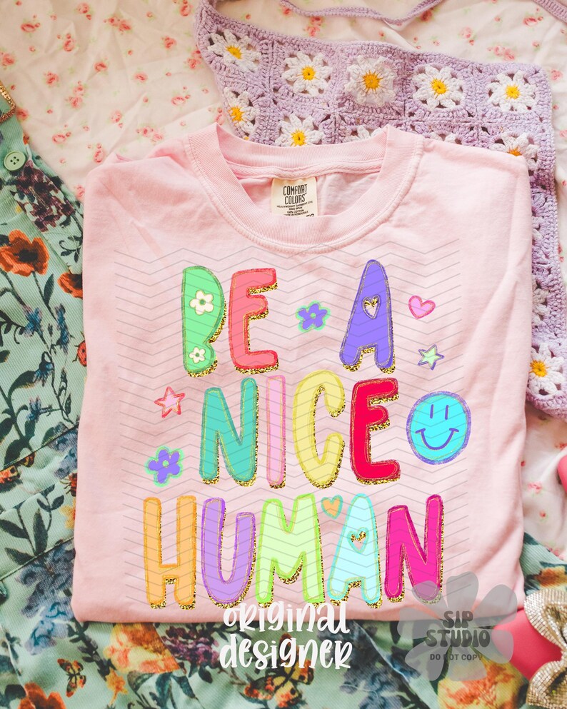 May include: A light pink Comfort Colors t-shirt with the phrase "BE A NICE HUMAN" in colorful, glitter-outlined letters. The design includes stars, flowers, and a smiley face. The words "original designer" and "SIP STUDIO DO NOT COPY" are also printed on the shirt.