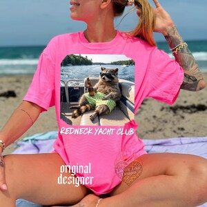 May include: A bright pink t-shirt featuring a graphic of a raccoon wearing sunglasses and green striped shorts, holding a beer bottle on a boat. The text "REDNECK YACHT CLUB" is below the graphic.