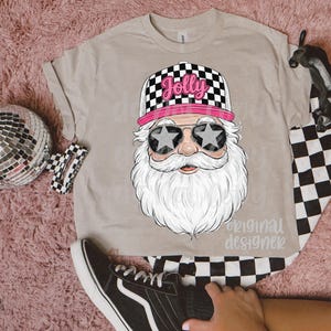 May include: A beige t-shirt featuring a graphic of Santa Claus wearing star-shaped sunglasses and a checkered hat with the word "Jolly". The design includes a long white beard and a pink brim on the hat. The text "original designer" is also visible.