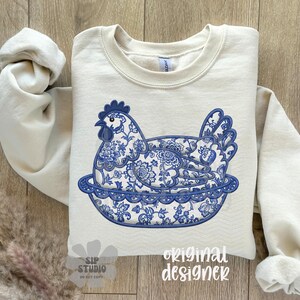 May include: A cream-colored sweatshirt featuring a blue and white floral design of a hen in a nest. The design is reminiscent of porcelain patterns. The words "original designer" are printed on the bottom right.