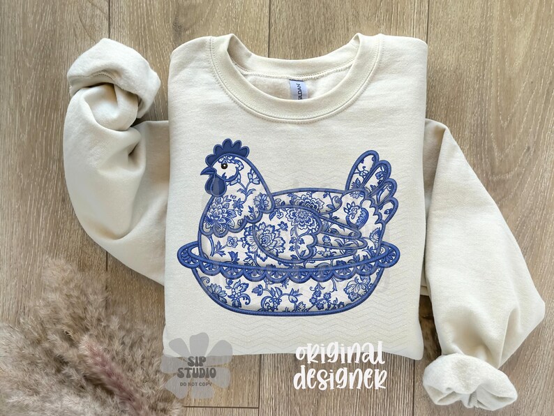 May include: A cream-colored sweatshirt featuring a blue and white floral design of a hen in a nest. The design is reminiscent of porcelain patterns. The words "original designer" are printed on the bottom right.