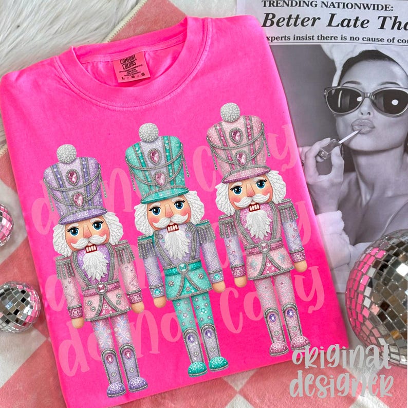 May include: A vibrant pink sweatshirt with three pastel-colored nutcracker designs. The nutcrackers are adorned with detailed hats and uniforms. The words "danon copy" are printed on the shirt. The text "original designer" is also printed on the shirt.