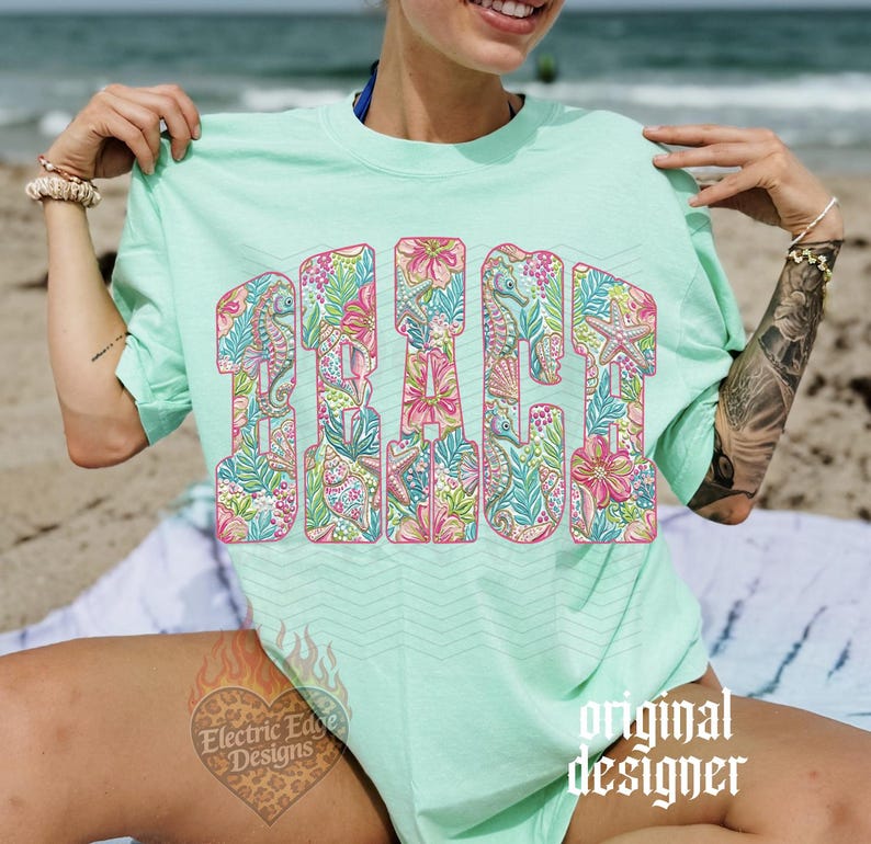 May include: A light green t-shirt with the word "BEACH" in large, colorful letters. The letters are filled with a floral and aquatic pattern, including seahorses and starfish. The shirt is a relaxed fit.